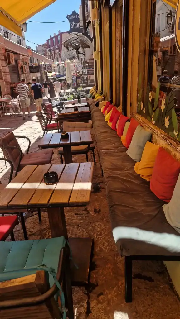 Outdoor seating at a cafe in Skopje's Old Bazaar, with wooden tables, cushioned chairs, and a long bench lined with colorful pillows beneath a yellow awning.