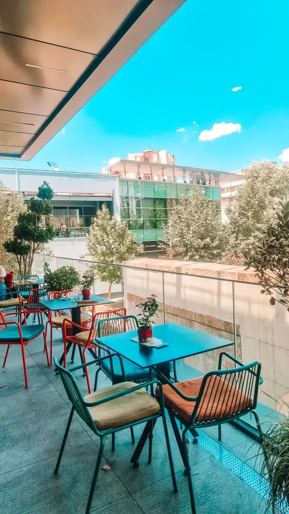 A modern terrace cafe in Skopje with vibrant red and green chairs, blue tabletops, and potted plants, overlooking a quiet street and green glass buildings.