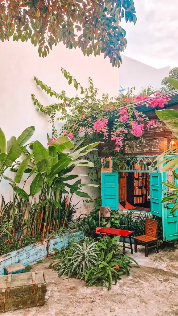 A vibrant garden nook outside a café with turquoise doors, lush tropical plants, and a bookshelf visible through the open window. Pink bougainvillea climbs over the rooftop, creating a dreamy reading and coffee spot.