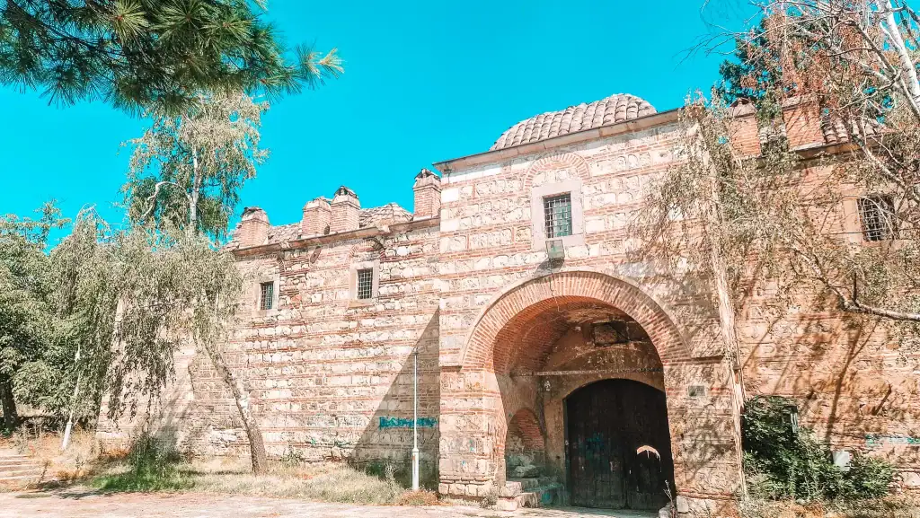 Exterior of an old Ottoman-style stone building with arched entrances and small barred windows, surrounded by trees and bathed in sunlight.