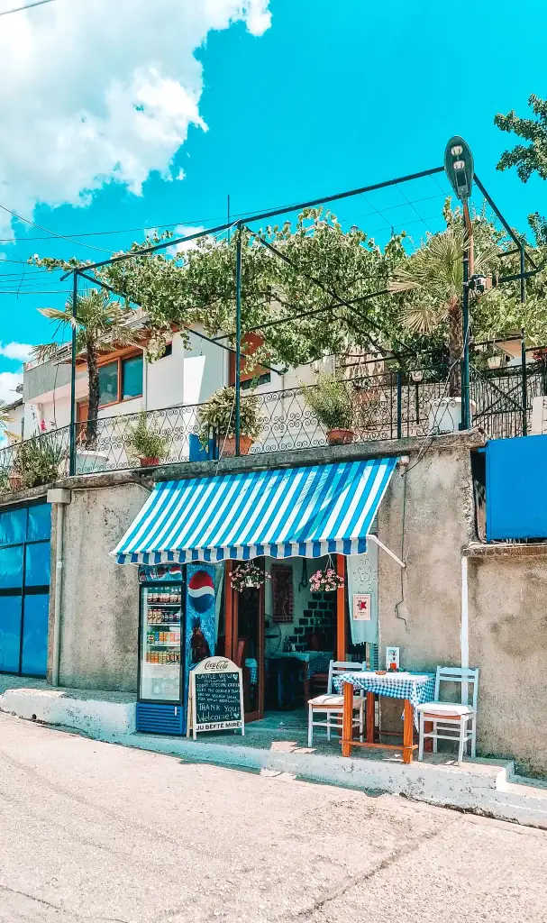 A quaint roadside café in Gjirokaster with a blue-and-white striped awning, small outdoor seating with checkered tablecloths, and a chalkboard welcome sign offering refreshments to visitors of the nearby castle.