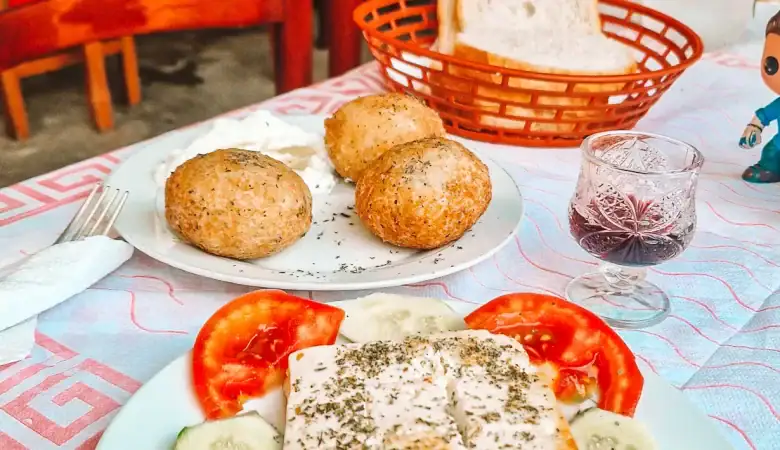 A traditional Albanian meal on a table with a pink patterned cloth, featuring grilled cheese with herbs, tomato and cucumber slices, a plate of three baked qifqi rice balls, slices of white bread, and a glass of red wine.