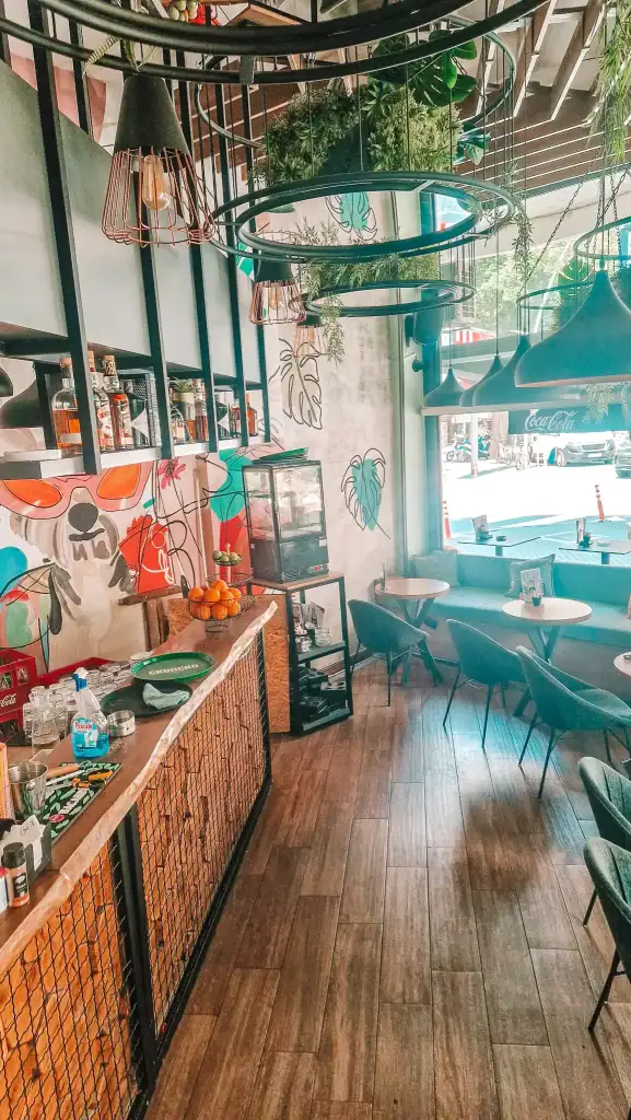 Interior of a stylish Skopje cafe with wooden floors, a curved bar, artistic wall murals, and large industrial lights hanging above cozy seating areas.