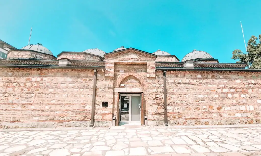 Front view of a historic hammam-turned-museum in Skopje with a symmetrical brick facade, arched doorway, and domed rooftops against a bright blue sky.