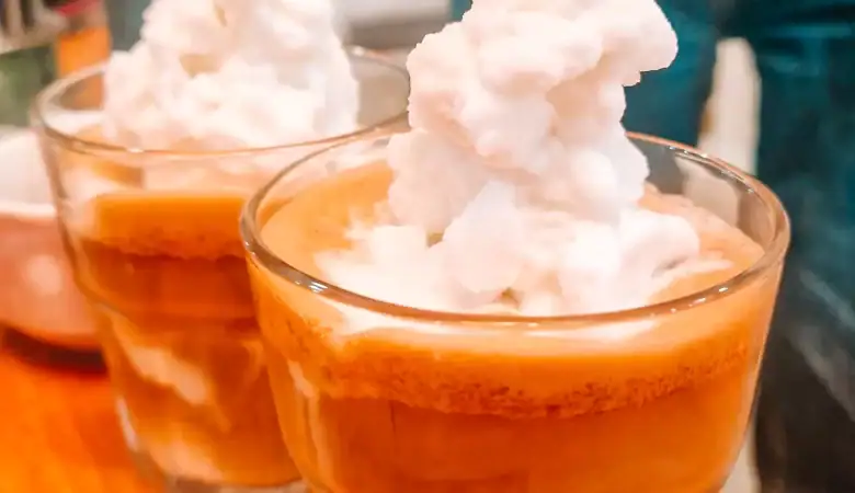 Two short glasses of coconut coffee topped with fluffy whipped cream, sitting on a wooden table inside a Vietnamese café. The drink's creamy layers are distinctly visible through the clear glasses.