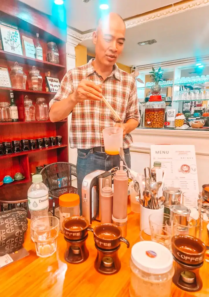 A barista stirs a drink behind a coffee bar lined with brewing tools, glass jars, and a detailed menu. The cozy interior with wooden shelves adds to the warm, educational feel of this Vietnamese coffee guide setting.