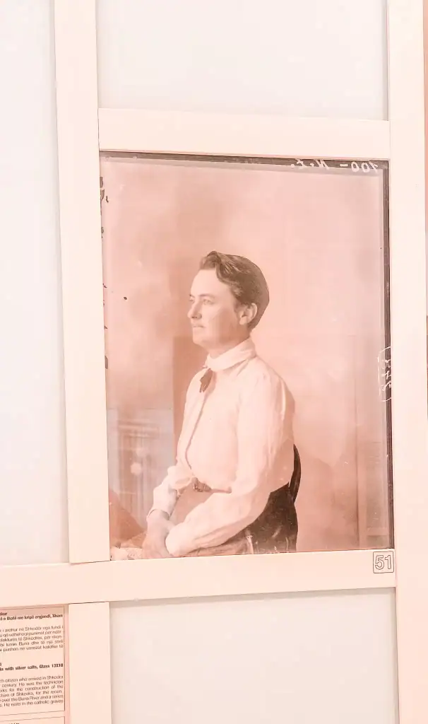 A sepia-toned photograph of a poised woman in early 20th-century attire displayed in a museum, framed with glass and surrounded by white panels and explanatory text.