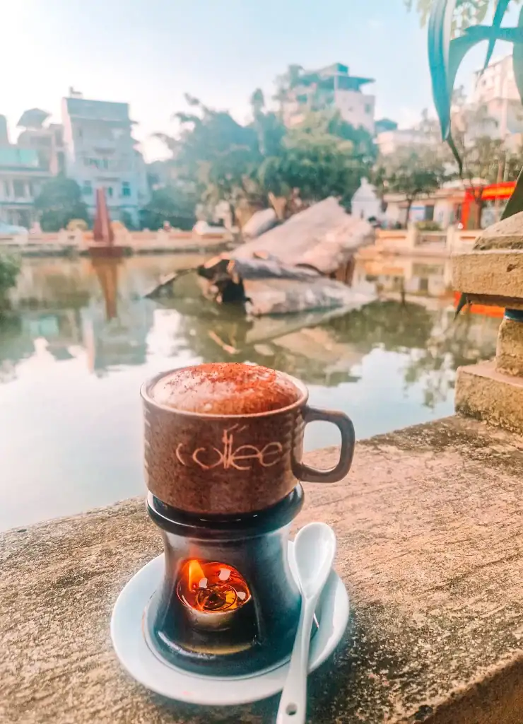 A hot cup of Vietnamese coffee is kept warm over a tea light candle beside a peaceful pond with a crashed airplane monument. The rustic ceramic cup and lakeside backdrop reflect a serene coffee moment.