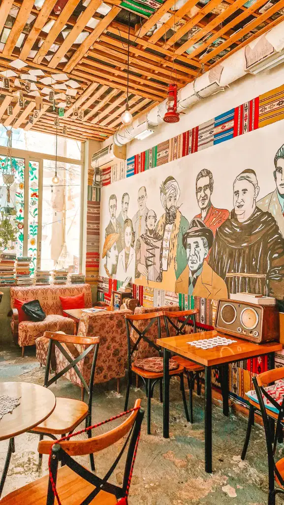 Eclectically decorated café in Shkoder featuring a vibrant mural of historical and cultural figures, patterned fabric wall panels, wooden furniture, and vintage touches like an old radio.