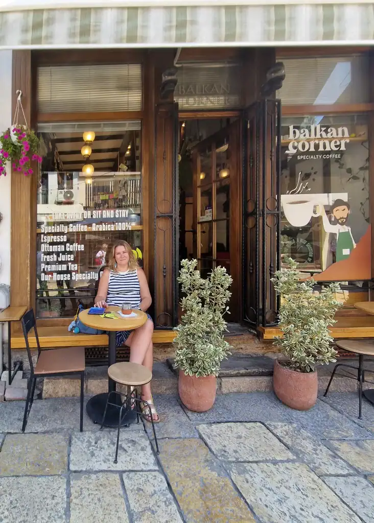 Em sitting outside Balkan Corner cafe in Skopje, sipping coffee at a small round table in front of a striped awning and glass doors with signs listing specialty drinks.