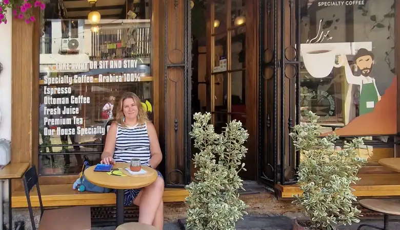 Em sitting outside Balkan Corner cafe in Skopje, sipping coffee at a small round table in front of a striped awning and glass doors with signs listing specialty drinks.