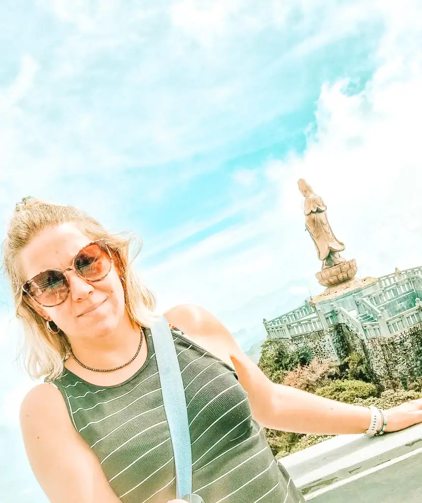 Em wearing sunglasses and a striped tank top stands in front of a large statue of the Buddhist goddess Guan Yin on Mount Fansipan, Vietnam, with bright blue skies and clouds in the background.