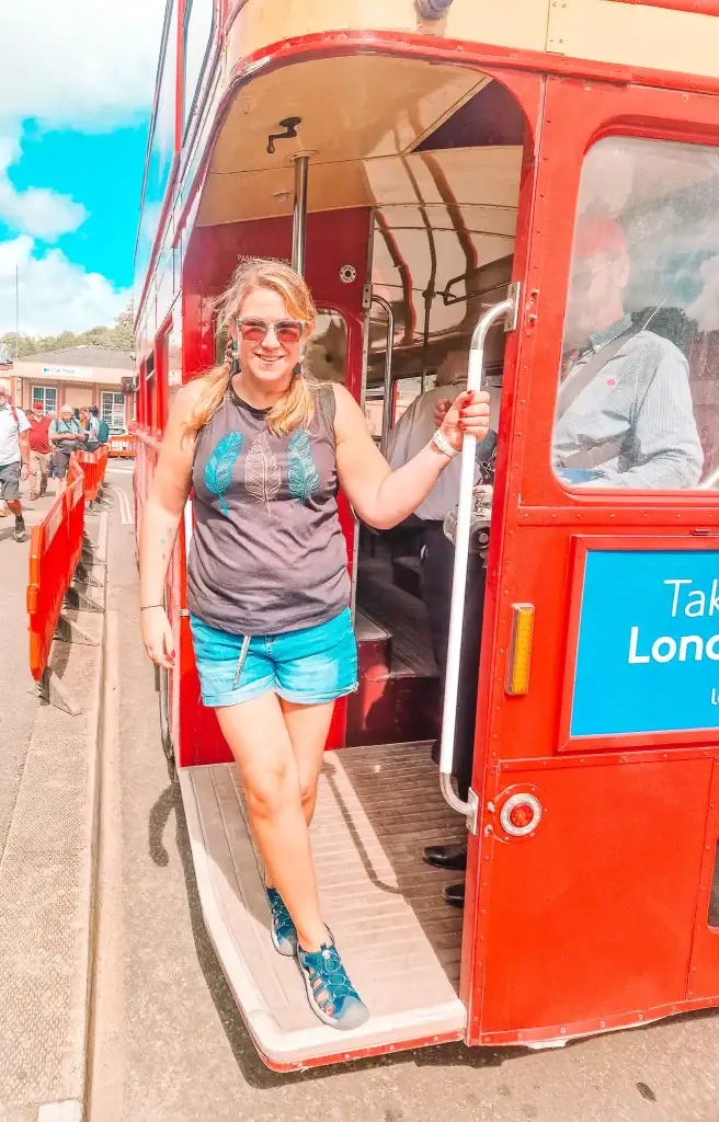 Em steps off a classic red London double-decker bus, smiling in casual clothes and sunglasses. The background shows other travelers and a car park, likely part of a tour experience.