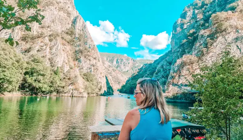 Em gazes across the calm green waters of Matka Canyon, surrounded by steep rocky cliffs under a clear blue sky. Kayakers and boats in the distance highlight this as a peaceful and scenic day trip from Skopje.