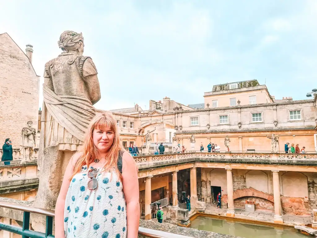 Em standing in front of a stone statue at the Roman Baths in Bath, with the ancient courtyard and green thermal pool visible in the background.