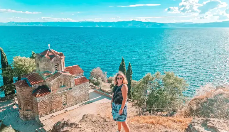 Em standing on a cliffside path with the Church of St. John at Kaneo and Lake Ohrid stretching out behind her under a sunny sky.