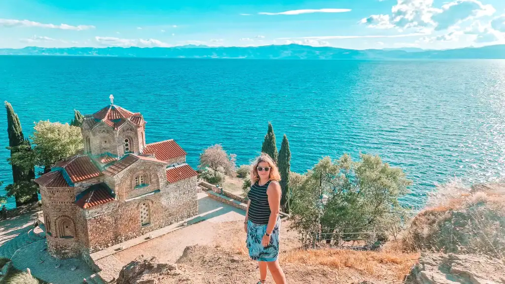 Em seated under a stone arch in Perast, Montenegro, with Our Lady of the Rocks island and the Bay of Kotor behind her, highlighting Montenegro’s coastal charm and mediaeval architecture.