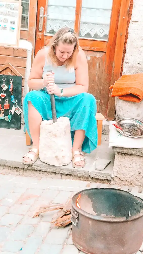 Em uses a traditional stone mortar and pestle outside a local artisan shop in Gjirokaster. Keychains and handmade souvenirs are displayed nearby, with a fire-heated cooking pot in the foreground.