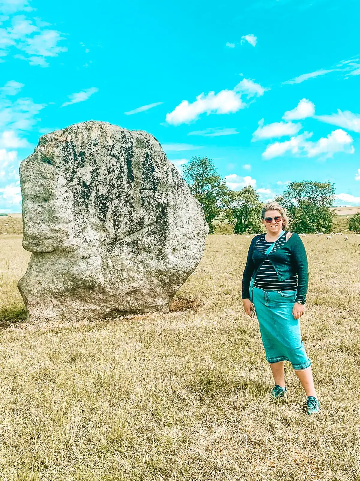 Em stands smiling in front of a large standing stone in an open grassy field under a bright blue sky. This photo highlights one of the iconic Neolithic monuments in Wiltshire.