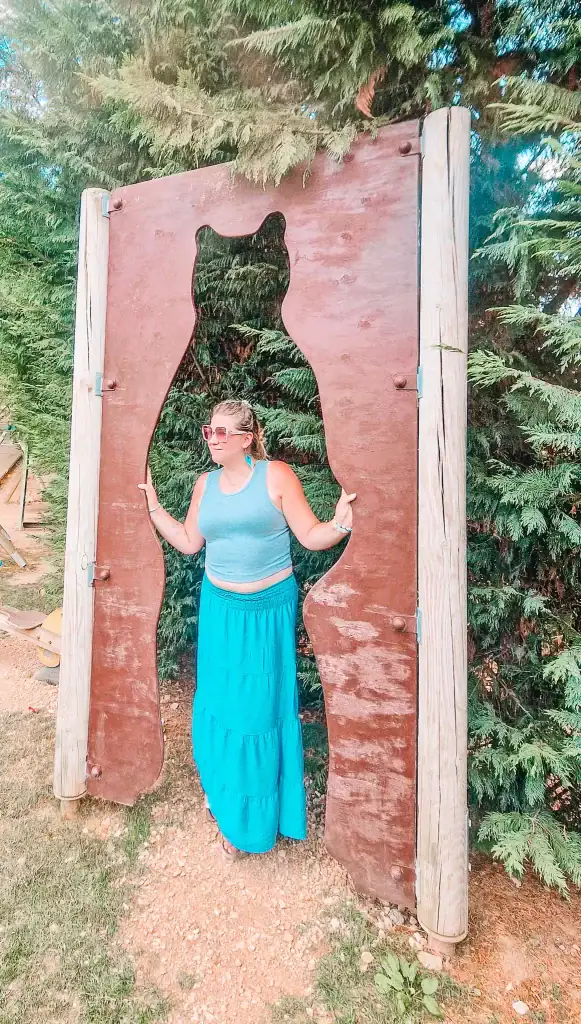 Em stands inside a large wooden cutout shaped like a bear silhouette, framed by tall evergreens. She smiles and holds the sides of the cutout while posing for a photo.