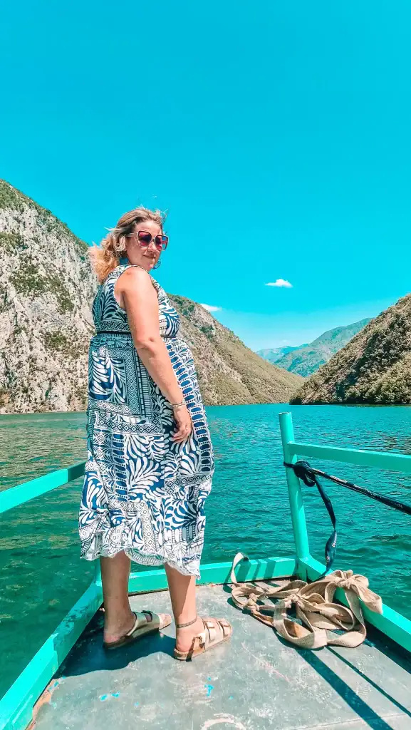 With wind in her hair and a bold patterned dress, Em stands confidently at the front of a turquoise boat, surrounded by lush mountain views and sparkling lake water.
