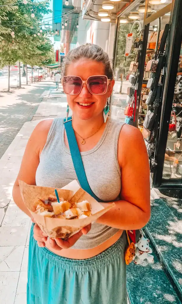 Em smiles while holding a tray of local street food on a sunny sidewalk lined with shops and outdoor string lights. She's wearing large sunglasses, a grey tank top, and a teal skirt.