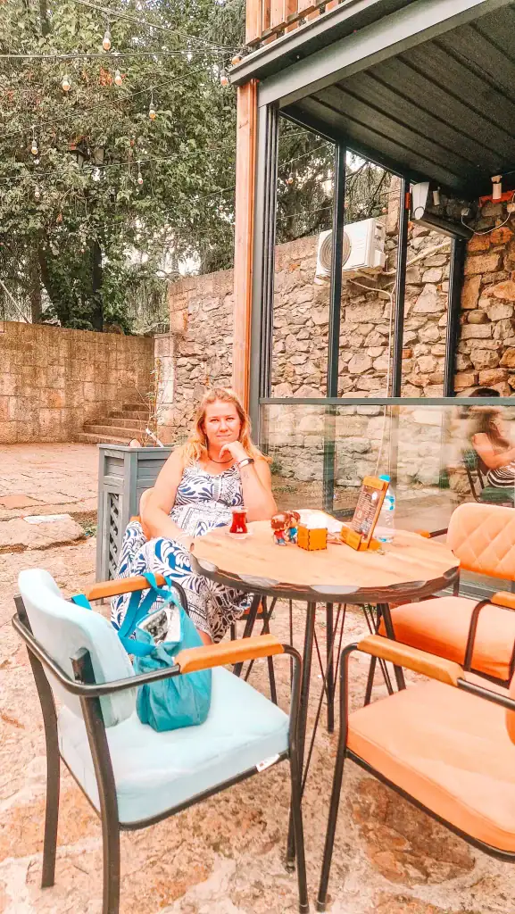 Em sits at an outdoor café in Skopje, enjoying tea on a sunny patio with stone walls, string lights overhead, and a relaxed atmosphere.