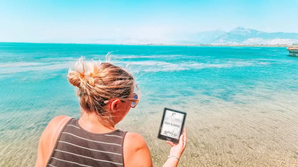 Em holds a Kindle displaying the cover of High Albania by Edith Durham while standing in shallow crystal-clear water, with mountain silhouettes in the distance under a bright blue sky.