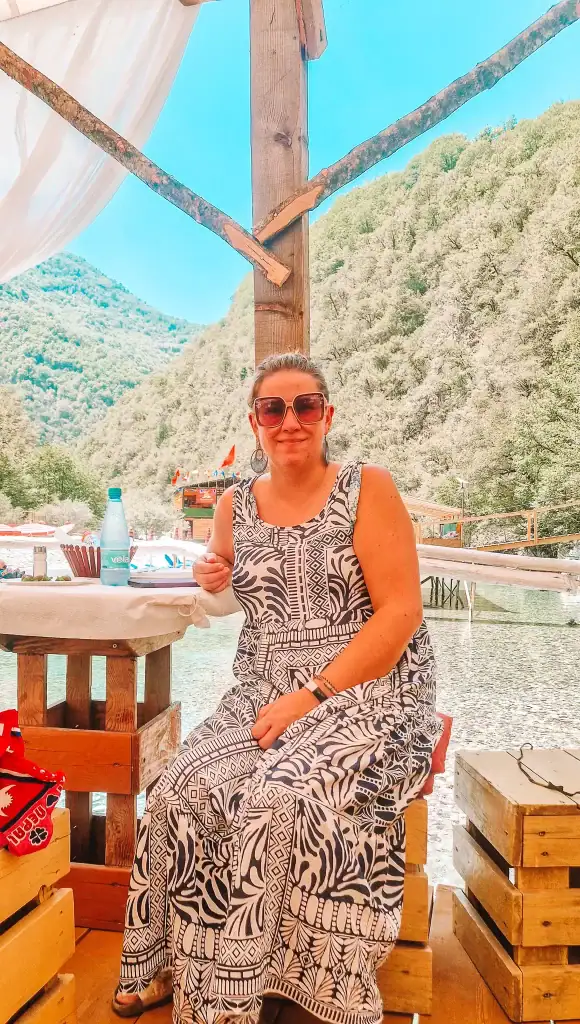 Em smiles while sitting at a rustic wooden table by a clear mountain stream, surrounded by wooden décor and shaded by white curtains in a peaceful outdoor setting.
