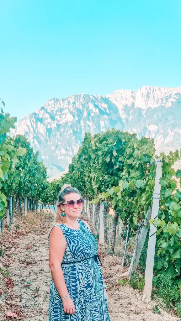 Em standing between grapevine rows in a vineyard, with clusters of green grapes visible and dramatic mountains behind her.
