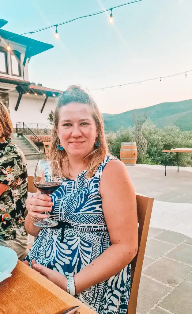 Em smiling and holding a glass of red wine at an outdoor table during a wine tasting, with mountains and a winery terrace in the background.