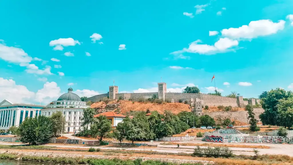 A hilltop view of Skopje Fortress, with its stone towers and Macedonian flag flying high above terracotta-roofed buildings and graffiti-covered walls below.