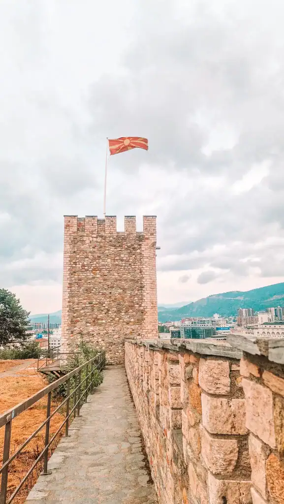 A close-up of a tower at Skopje Fortress, with the Macedonian flag waving atop and panoramic views of the city and mountains stretching into the distance under a cloudy sky.