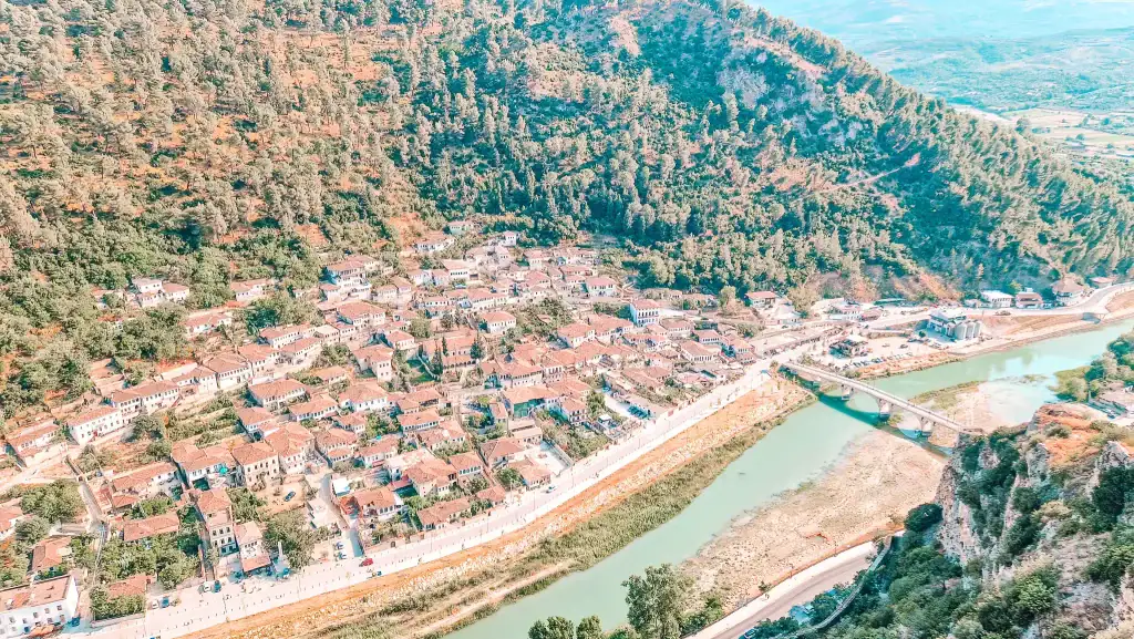 Panoramic aerial shot of Berat’s hillside covered in red-roofed Ottoman houses beside the Osum River, with a green forested mountain rising steeply behind.