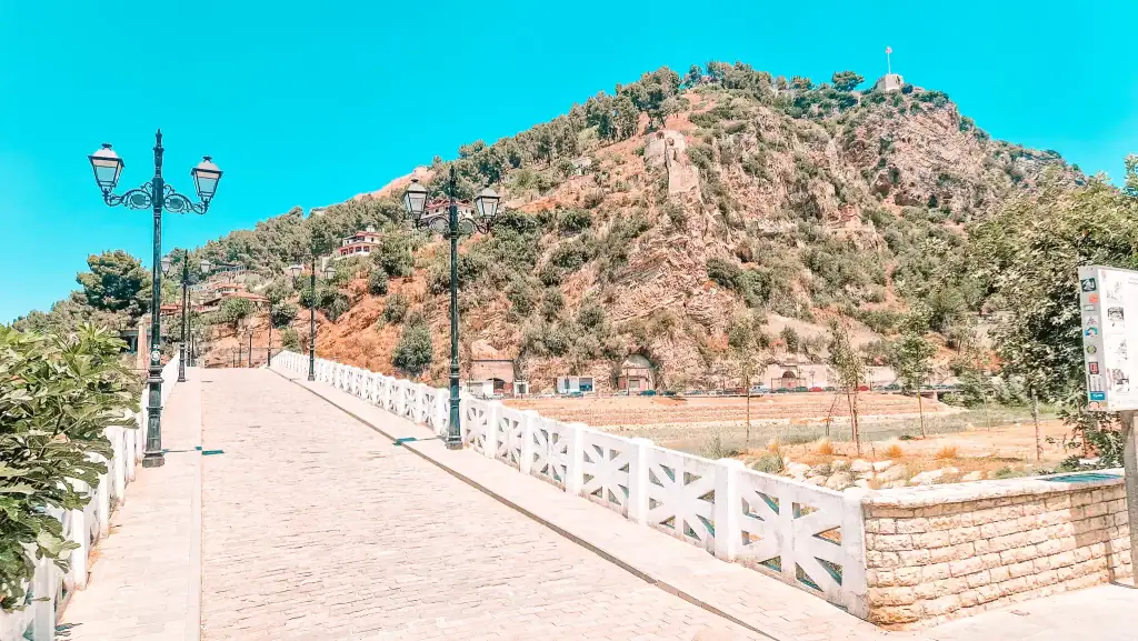 Wide cobblestone bridge with ornate lampposts crossing toward a rocky hill topped with a flag at Berat Castle.