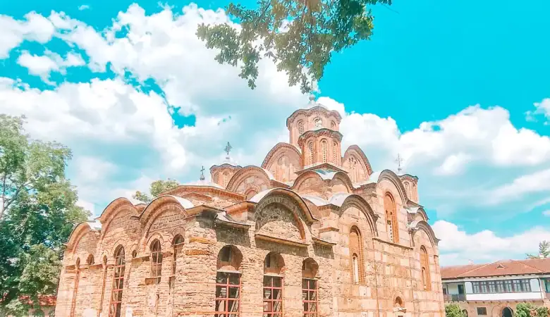 A historic Serbian Orthodox church with arched brickwork sits under a bright sky, framed by green grass and leafy trees, capturing religious heritage in the Balkans.