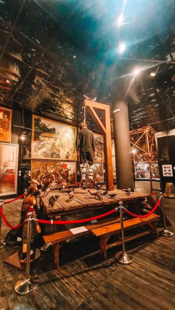 Museum exhibit depicting a life-sized execution scene with a hanging mannequin, surrounded by mannequins of children and pigeons, set against a backdrop of historical paintings.