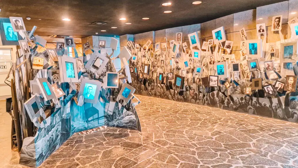Artistic museum installation with hundreds of black-and-white portrait photographs framed and lifted by illustrated hands on the wall, symbolizing remembrance and protest.