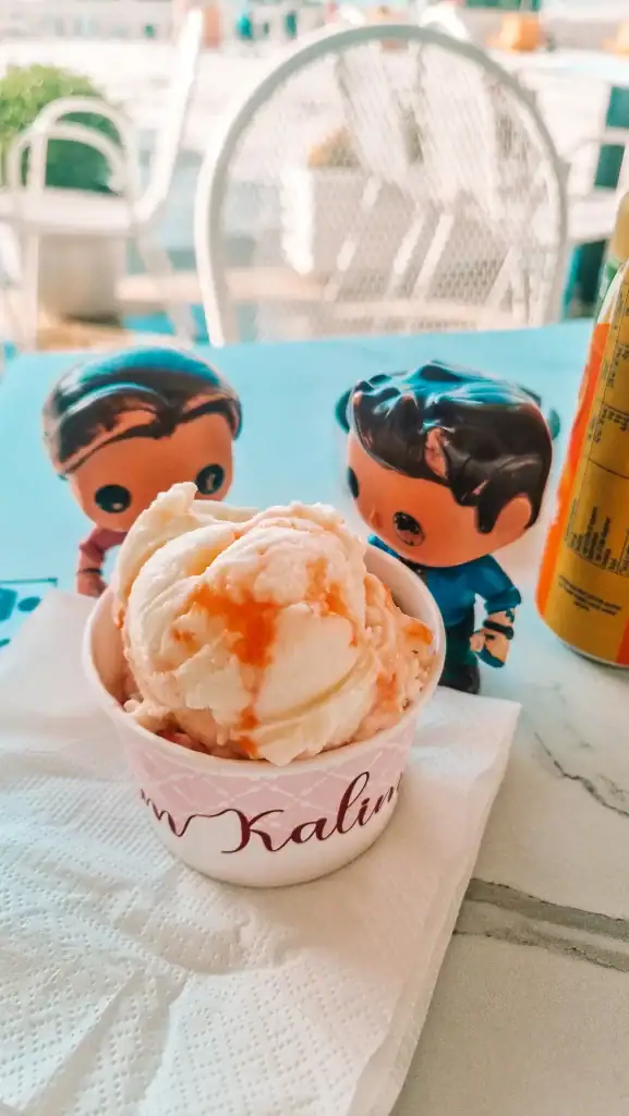 A small cup of vanilla ice cream with caramel drizzle sits on a table in Gjirokaster, flanked by two collectible toy figurines. The setting suggests a casual café stop during sightseeing.