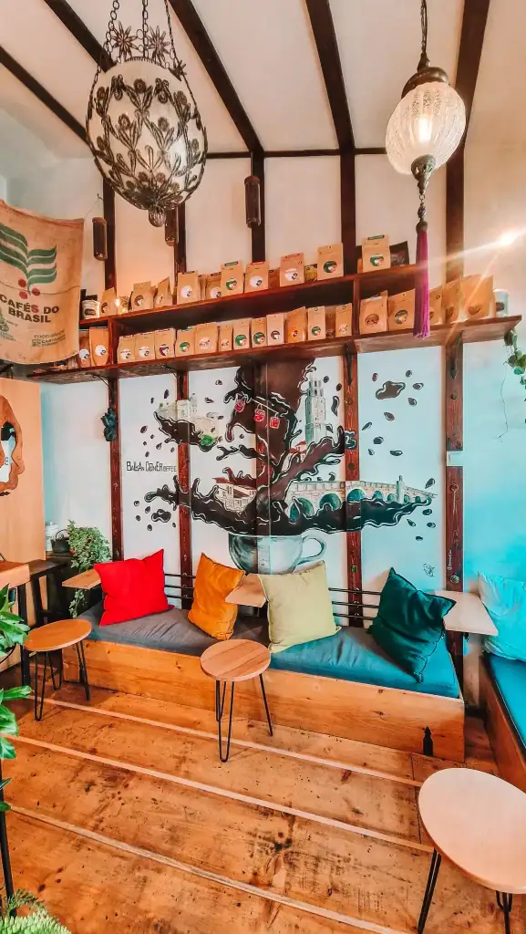The eclectic interior of Balkan Corner cafe featuring colorful pillows, wooden benches, and a large wall mural of coffee beans swirling into a cityscape.