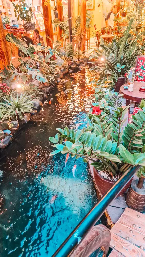 A narrow koi pond runs through the center of a lush, plant-filled café space with glowing warm lights. Diners sit beside the water surrounded by greenery and wooden decor, blending nature and coffee culture.
