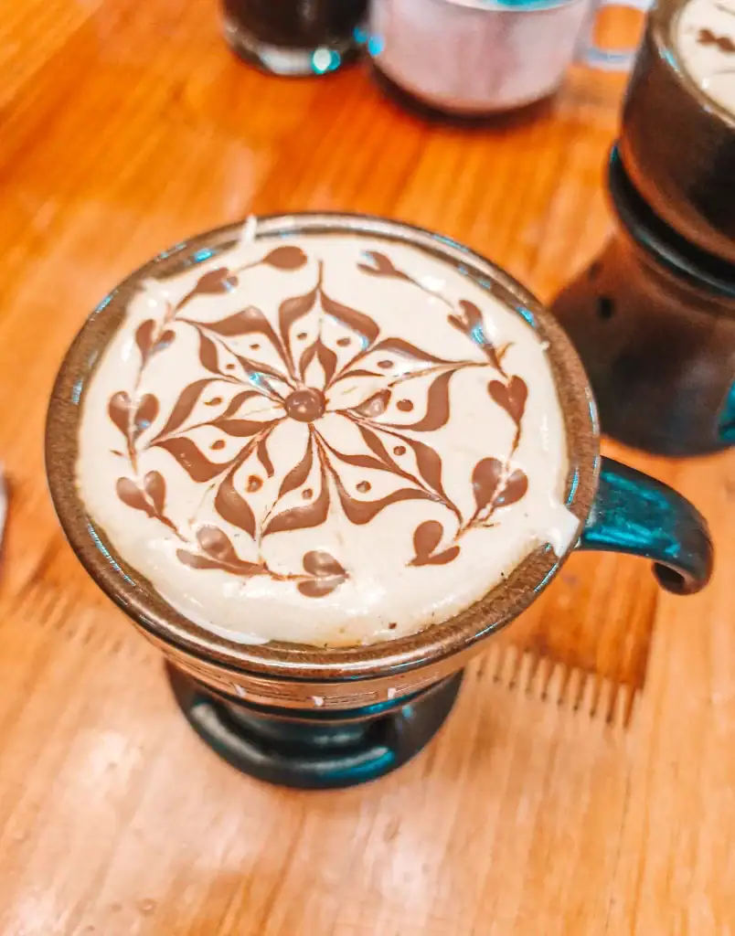 A close-up of Vietnamese egg coffee topped with artistic chocolate syrup in a flower pattern on the creamy foam. The drink is served in a ceramic cup on a wooden table, showcasing the craft behind this traditional beverage.