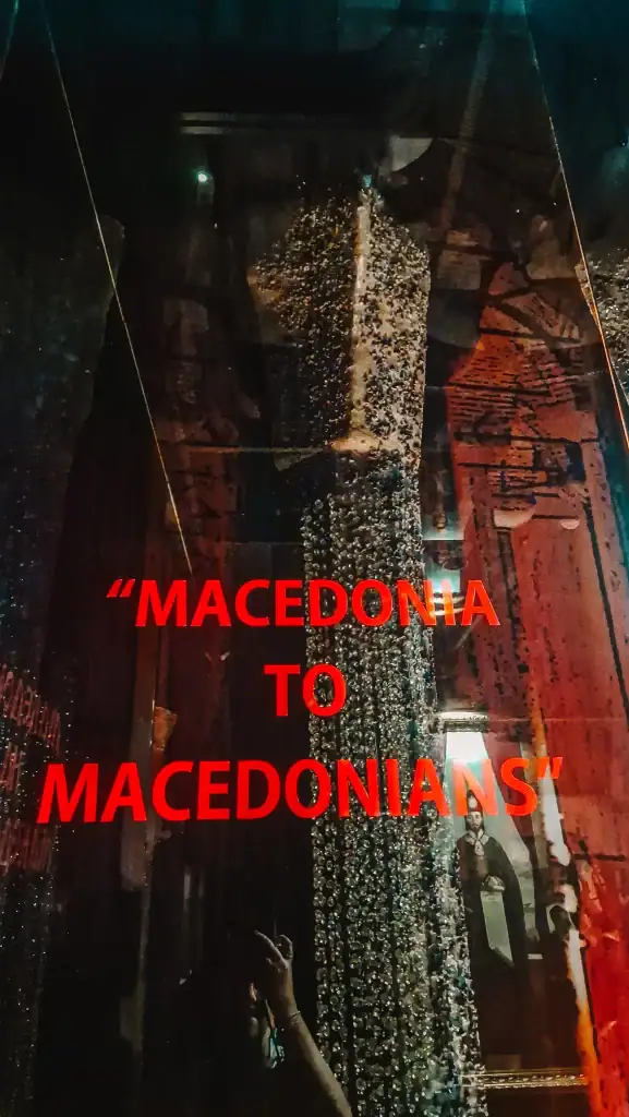Red text on glass reading “MACEDONIA TO MACEDONIANS” in a museum exhibit, with reflective lighting and a beaded installation behind it.