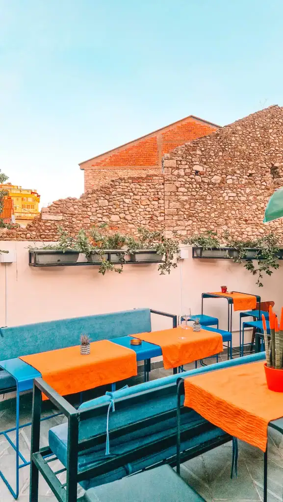 A colourful terrace café in Shkoder with bright orange tablecloths, sleek blue benches, and a stone wall backdrop under a clear blue sky.