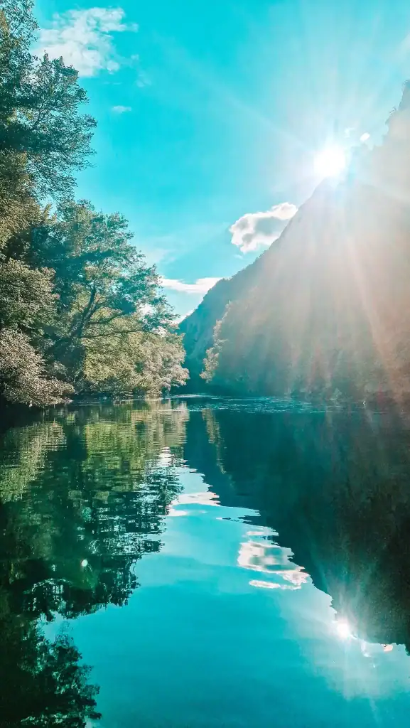 The sun beams over Matka Canyon, casting a soft glow through the mountain ridge and reflecting off the calm, glassy water surrounded by lush greenery.