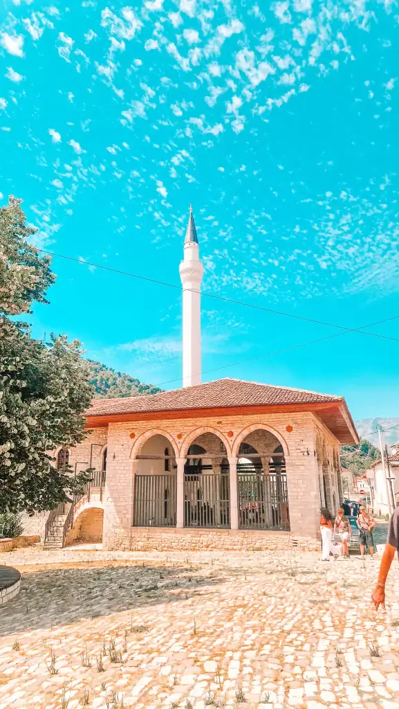 Stone mosque with arched portico and tall white minaret reaching into a bright blue sky, known as the King’s Mosque in Berat.