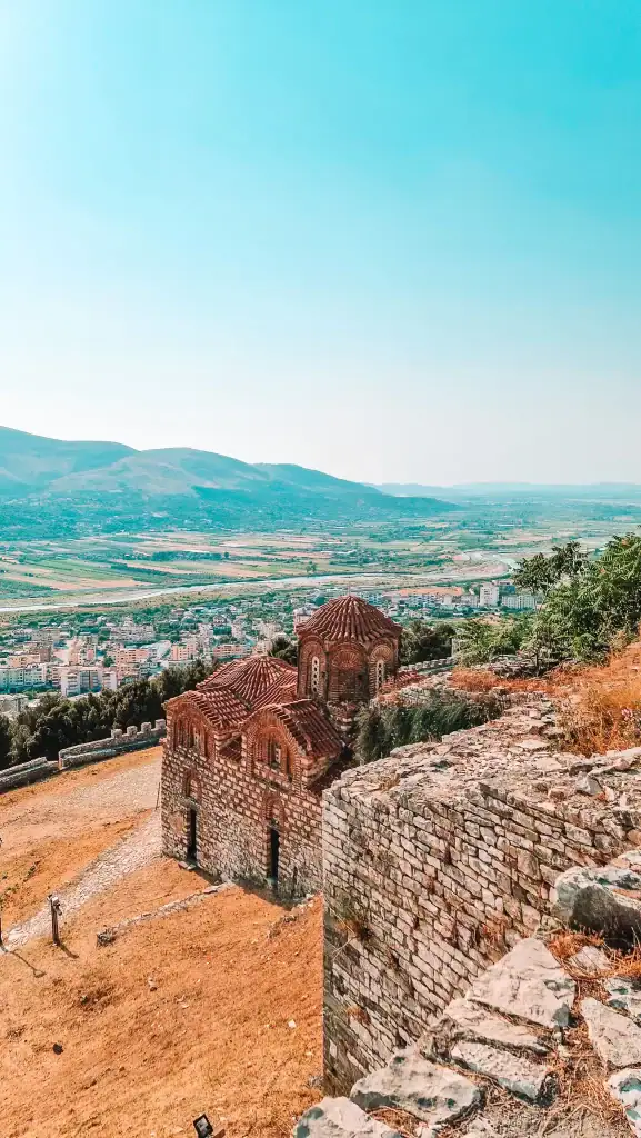 Small stone Byzantine church with red-tiled domes perched on a hill inside Berat Castle, overlooking the city and surrounding valley.