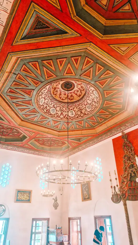 Intricately painted wooden ceiling with geometric and floral patterns inside the King’s Mosque, with chandeliers hanging below.