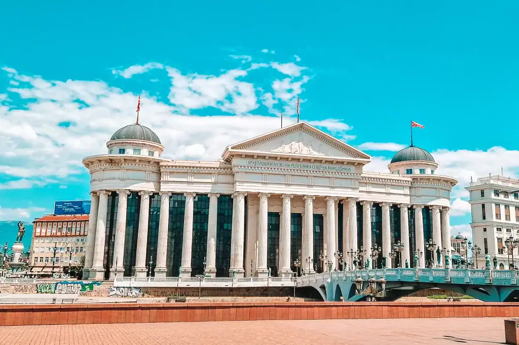 The Archaeological Museum of North Macedonia in Skopje features grand neoclassical architecture with tall white columns, domed roofs and flags flying atop.