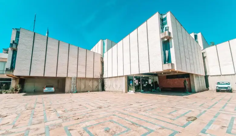 Wide shot of a modernist museum building in Skopje with geometric architecture, a tiled courtyard, and a few parked cars.